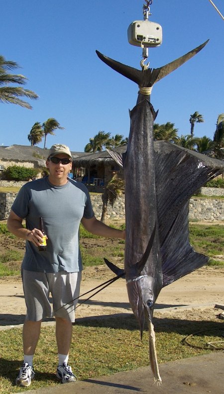 Man in sunglasses holding a beer beside a large sailfish hanging on a scale at a fishing dock