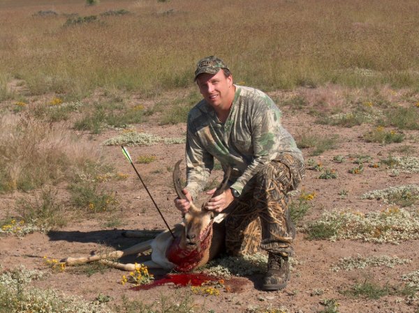 Hunter in camouflage kneeling beside a harvested pronghorn antelope taken with a bow in an open grassland field