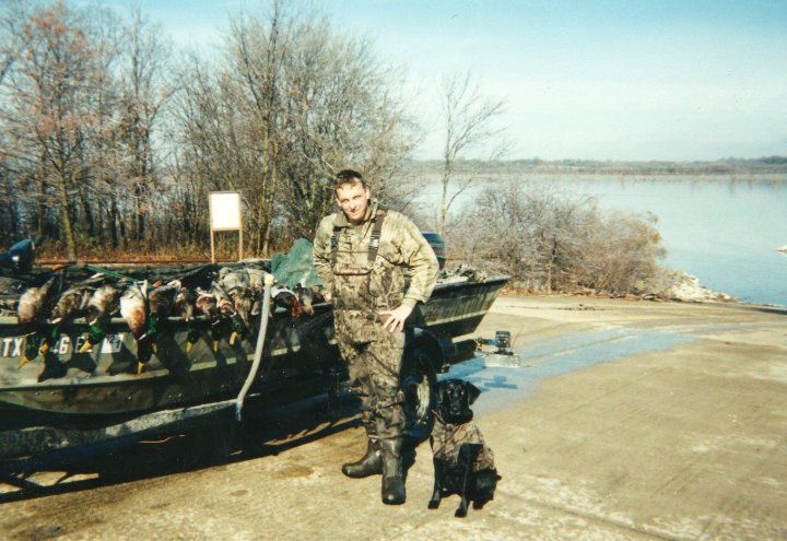 Hunter in camouflage standing by boat with duck harvest and black dog near lake shore