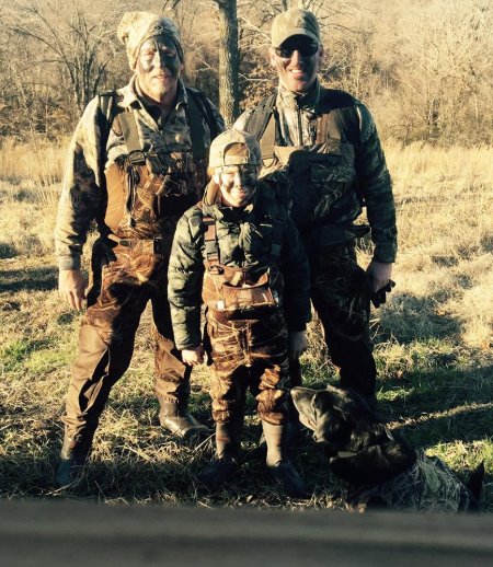 Two men and a boy in camouflage hunting gear standing with a hunting dog in a grassy field