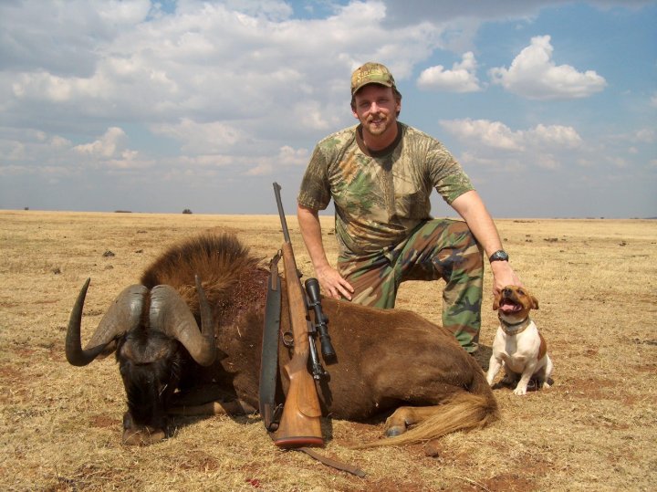 Hunter kneeling beside a black wildebeest trophy with rifle and dog on the African savanna