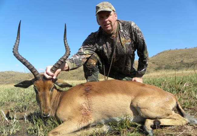 Hunter in camouflage kneeling beside a harvested impala with long spiral horns on grassy plains