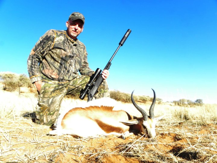 Hunter in camouflage kneeling beside a harvested springbok holding a rifle in the Kalahari