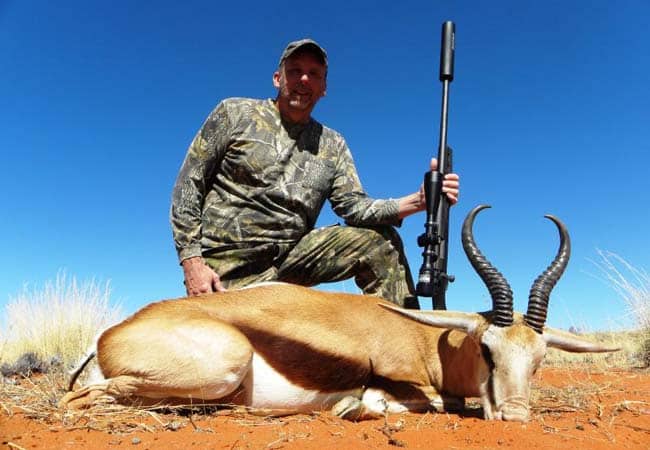 Hunter kneeling beside a springbok trophy holding a rifle under blue skies in Namibia