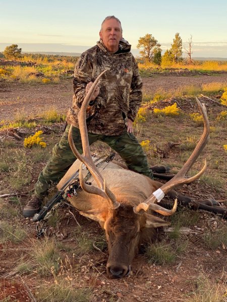 Hunter in camo poses behind a large trophy elk harvested in an open field at sunset