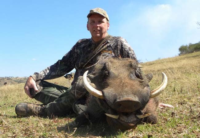 Hunter in camo kneeling beside a harvested warthog on a grassy African plain