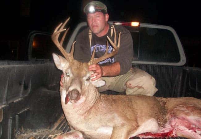 Hunter wearing headlamp kneels beside a large whitetail buck with antlers in truck bed at night
