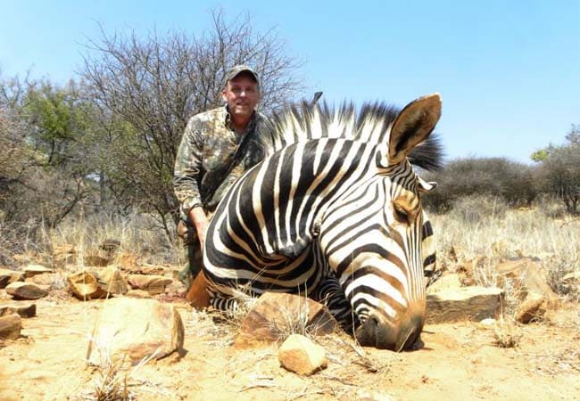 Hunter kneeling behind a zebra trophy in the South African bush under a clear blue sky