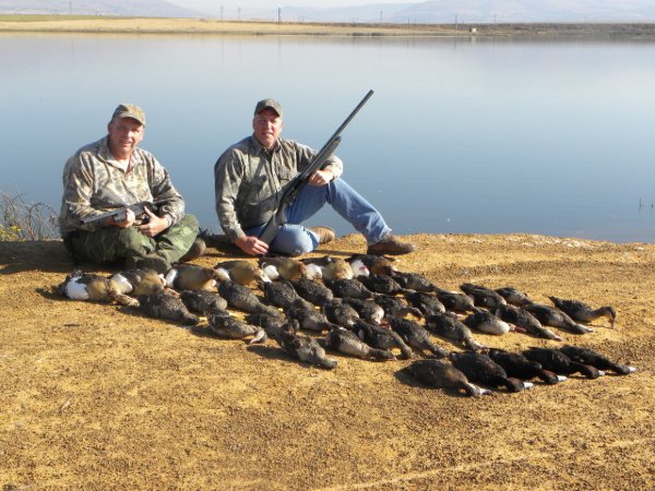Two hunters in camo sitting by a lake with a large harvest of ducks laid out in front of them