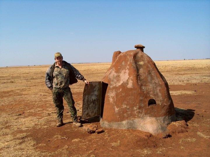 Man in camo standing beside a large old clay bread oven with metal door in a dry grassy field