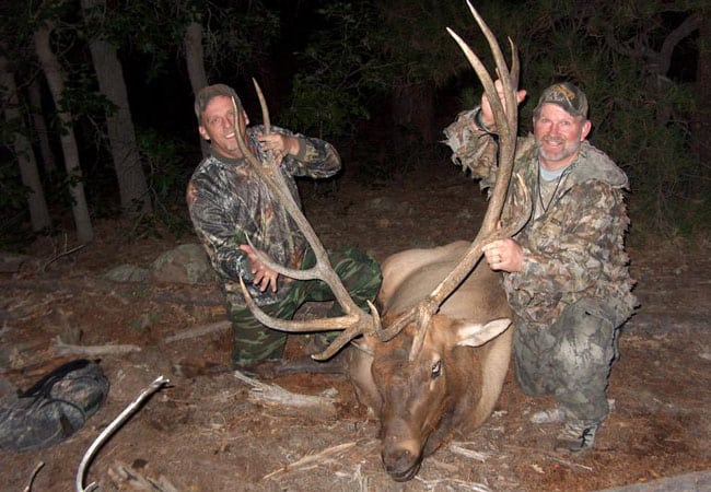 Two smiling hunters in camo kneeling beside a large bull elk with big antlers at night